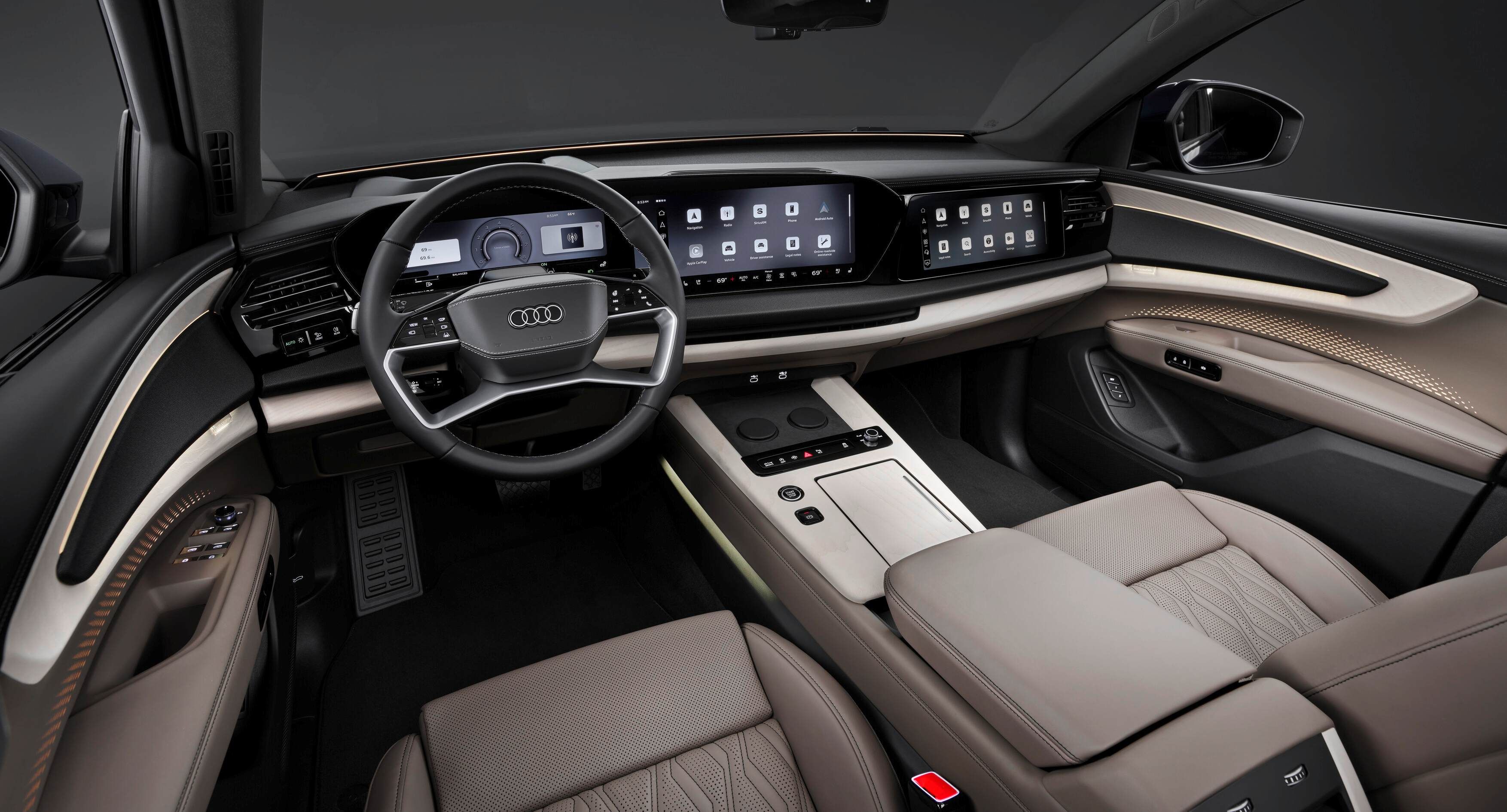Interior moderno do novo Audi Q9, visto do banco do motorista, com painel digital largo e telas sensíveis ao toque ocupando quase todo o console frontal. Os bancos são de couro bege, com acabamento sofisticado e iluminação ambiente discreta nas portas e no painel. O volante multifuncional, o console central largo e os poucos botões físicos reforçam o visual minimalista e tecnológico.