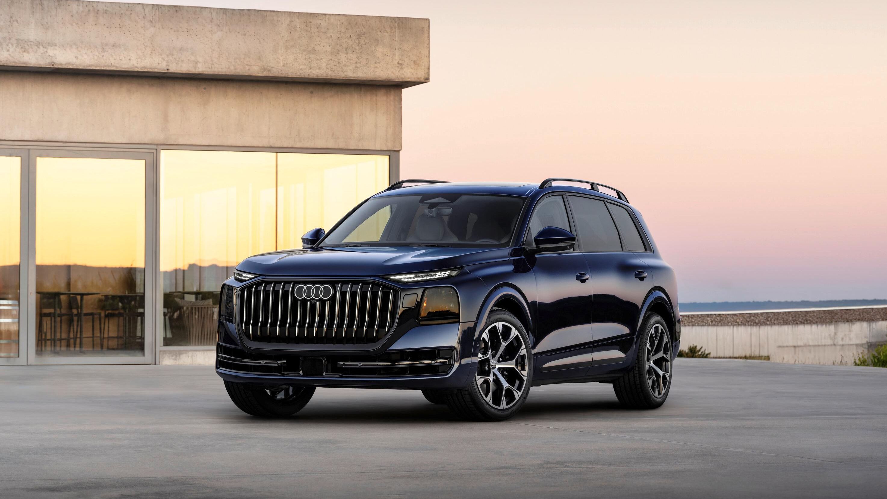 Novo Audi Q9 estacionado em uma esplanada ampla de concreto. Ao fundo, há uma casa de arquitetura minimalista com grandes janelas de vidro refletindo a luz do entardecer. O céu em tons suaves de rosa e azul cria um ambiente calmo e sofisticado.