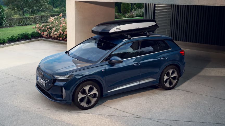 Der Q4 SUV e-tron mit einer Dachbox schräg von vorn auf einem Parkplatz eines modernen Hauses, im Hintergrund ein gepflegter Garten.