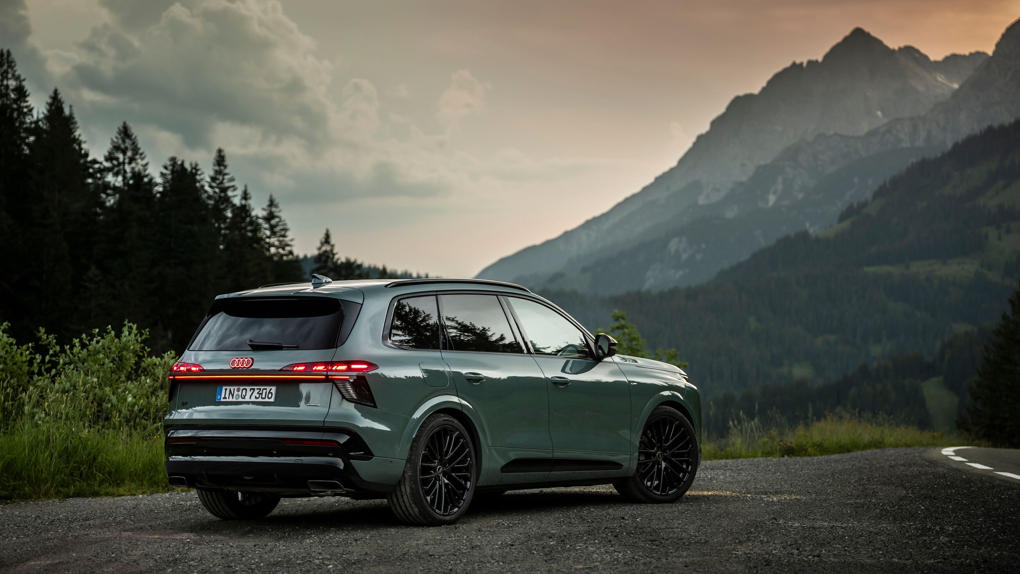 Novo Audi Q7, estacionado numa estrada de asfalto ao entardecer. Ao fundo, vê-se uma paisagem montanhosa com picos altos parcialmente cobertos por nuvens e cercados por floresta. 