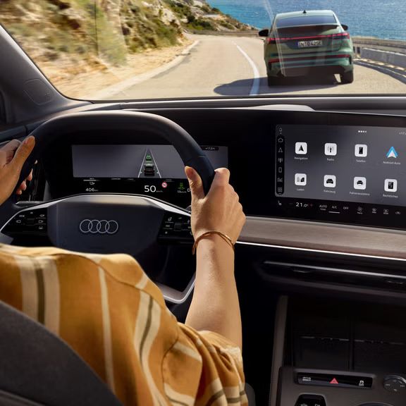 Blick von hinten auf das Cockpit des Q4 Sportback e-tron. Eine Frau sitzt am Lenkrad und steuert das Auto auf einer Bergstraße, zur rechten Wasser. Ein Fahrzeug fährt unmittelbar vor dem Q4 Sportback e-tron.