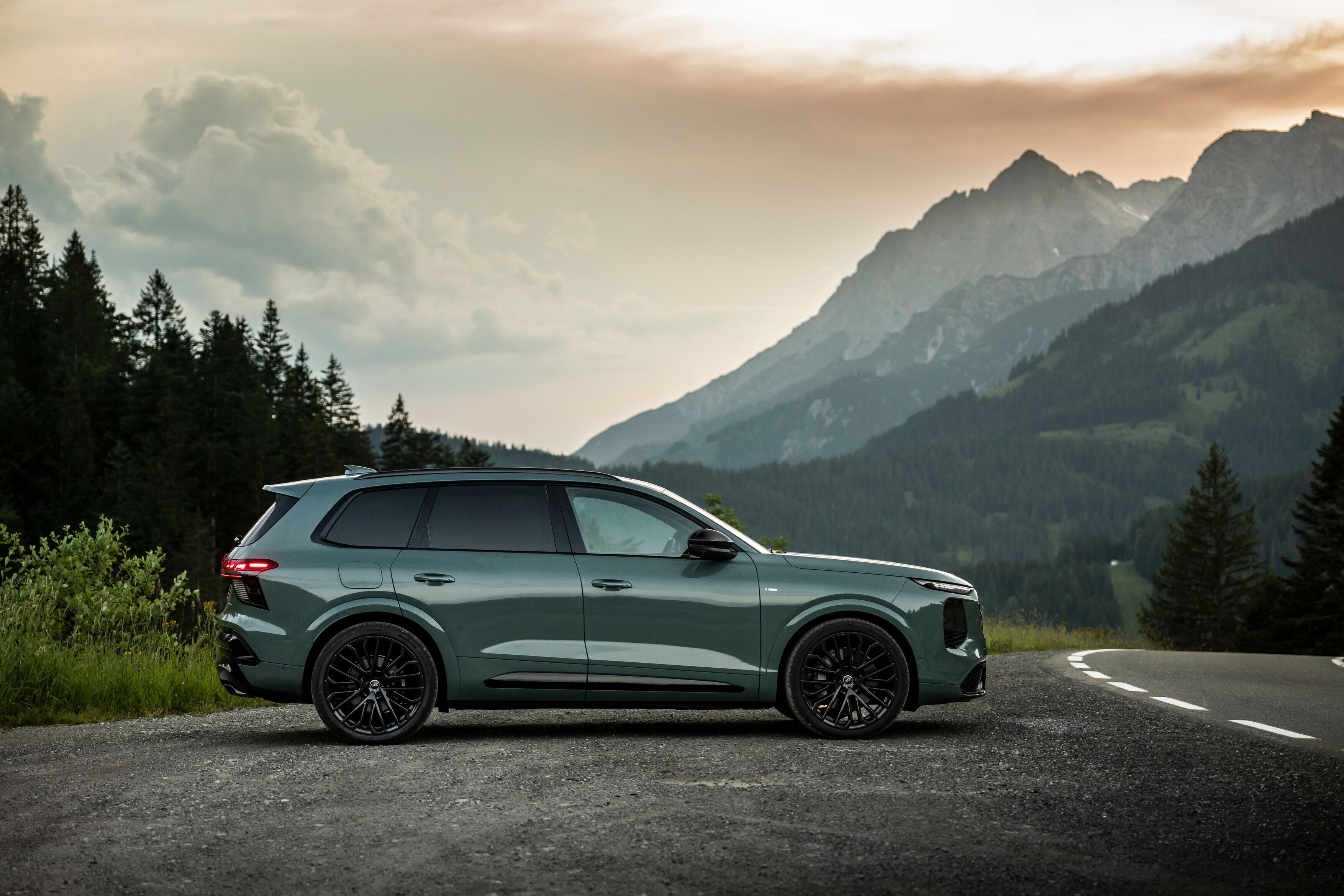 Novo Audi Q7 estacionado numa estrada de asfalto numa área montanhosa. Ao fundo, veem-se montanhas altas parcialmente cobertas por neblina e um céu de fim de tarde com nuvens iluminadas. 