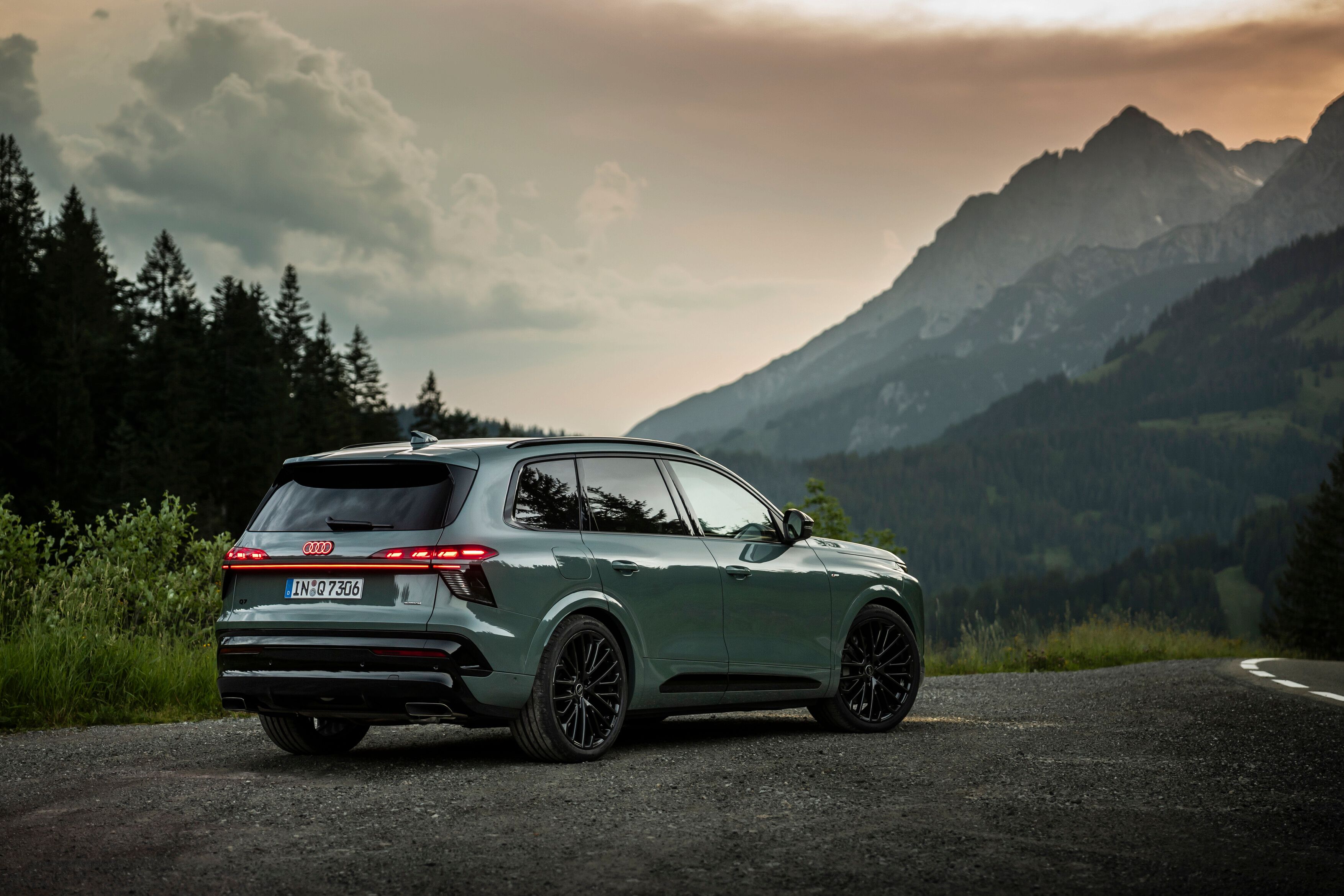 Audi Q7 novo, estacionado numa estrada de asfalto. Ao fundo, há montanhas altas parcialmente cobertas por nuvens e floresta densa de pinheiros.