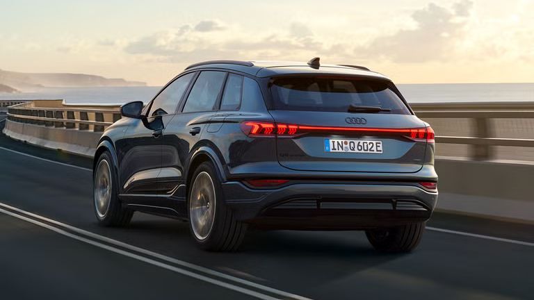 Foto traseira inclinada do SUV Q6 e-tron, que percorre uma estrada sinuosa à beira-mar. Montanhas podem ser vistas ao fundo.