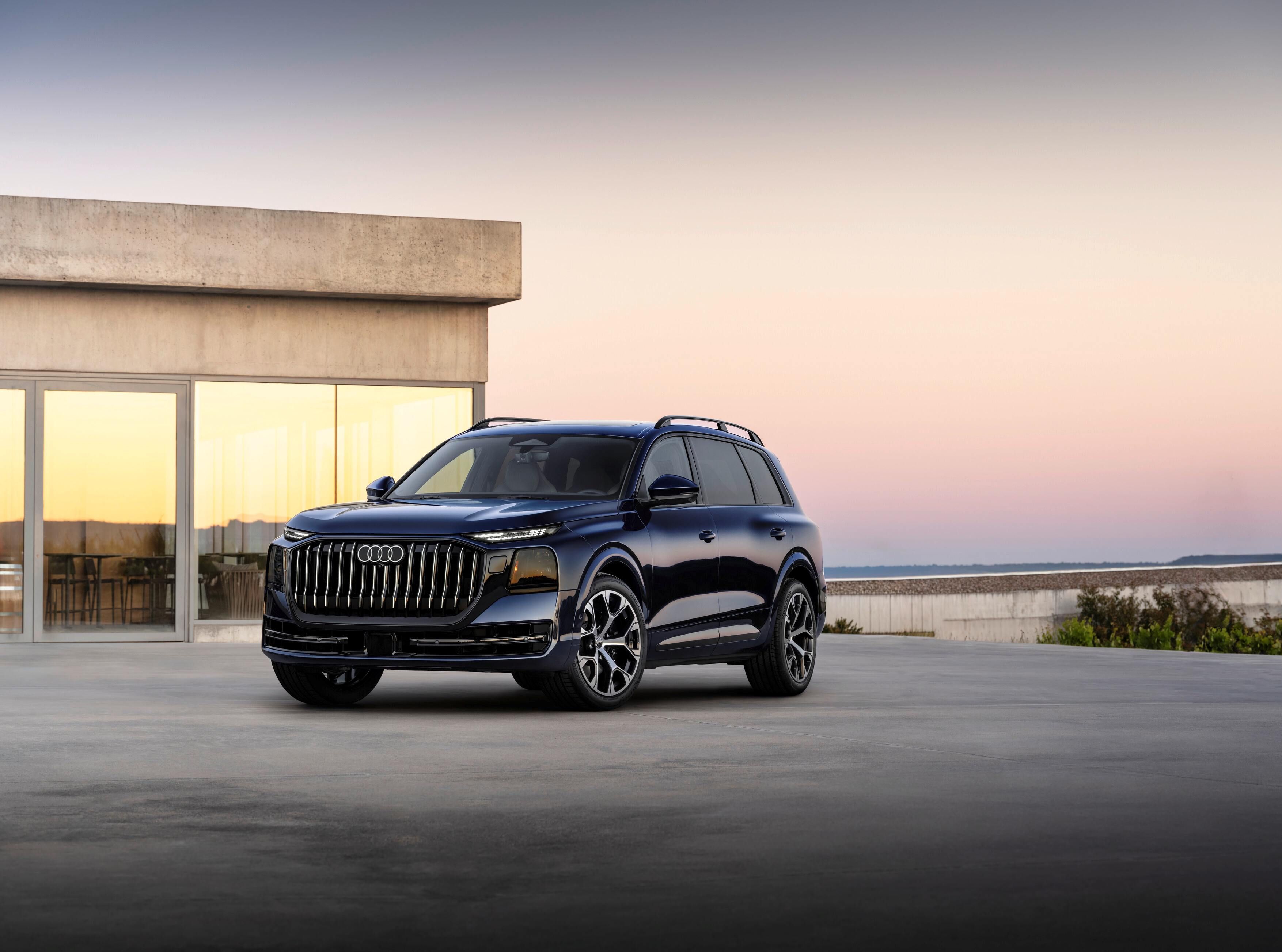Novo Audi Q9 estacionado em uma esplanada ampla de concreto. Ao fundo, há uma casa de arquitetura minimalista com grandes janelas de vidro refletindo a luz do entardecer. O céu em tons suaves de rosa e azul cria um ambiente calmo e sofisticado.