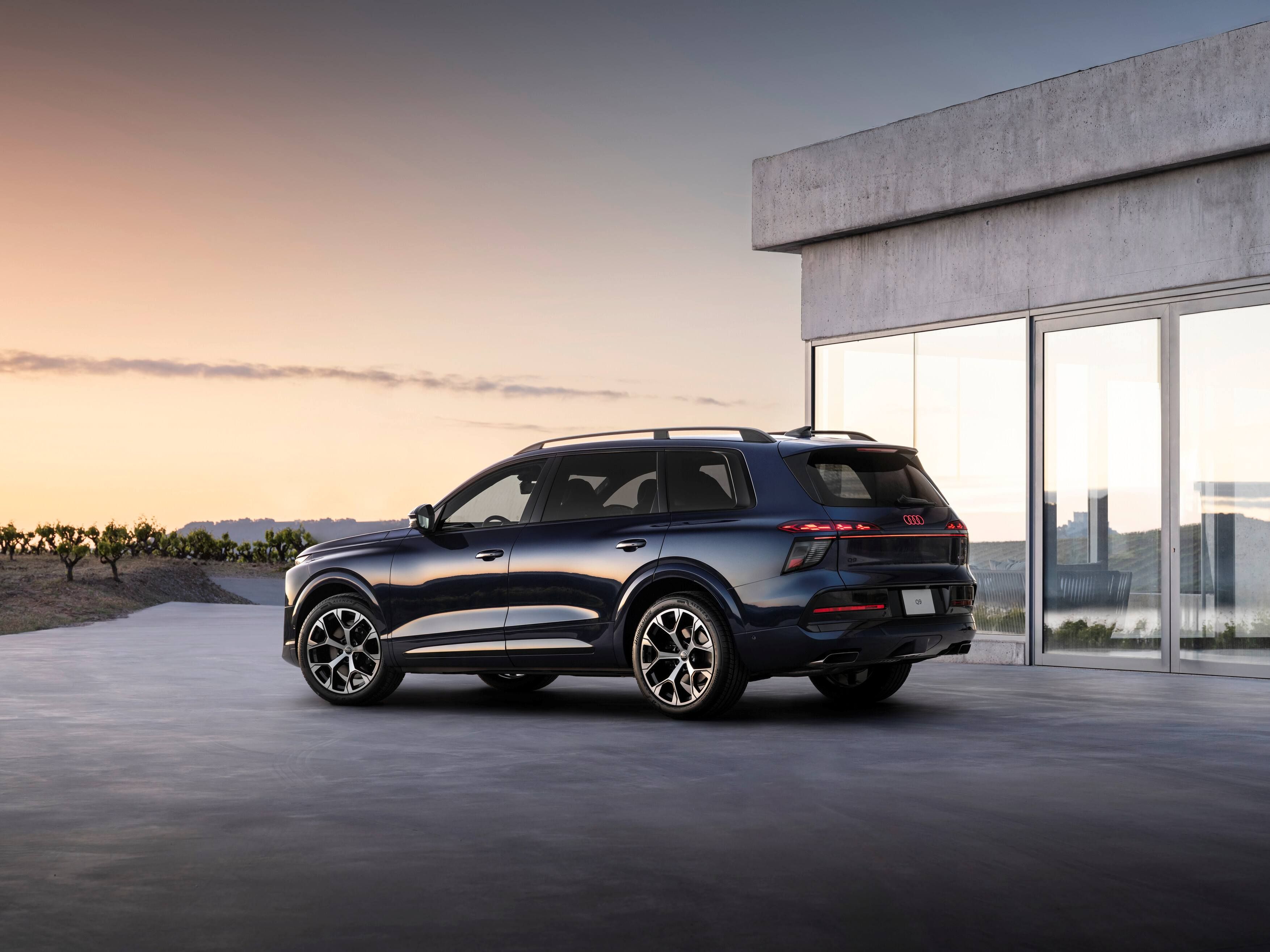 Novo Audi Q9 num tom azul-escuro metálico, estacionado em uma área aberta de concreto ao lado de uma casa de arquitetura minimalista com grandes painéis de vidro. O carro está visto em três quartos traseiros, com rodas grandes esportivas e lanternas traseiras em LED acesas. Ao fundo há um pôr do sol suave e uma plantação baixa, criando um ambiente elegante e tranquilo.