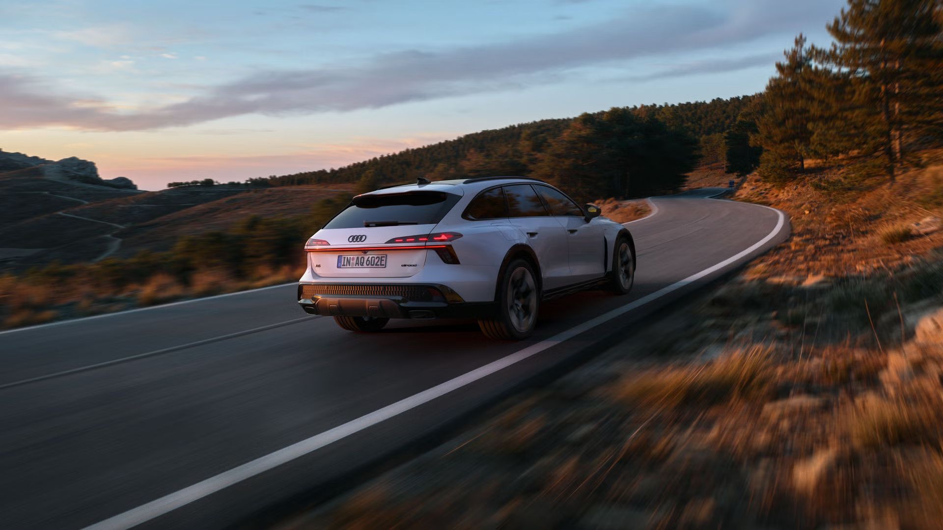 Der Audi A6 allroad e-hybrid schräg von hinten in voller Fahrt auf einer Gebirgsstraße. Im Hintergrund ein Waldstück.