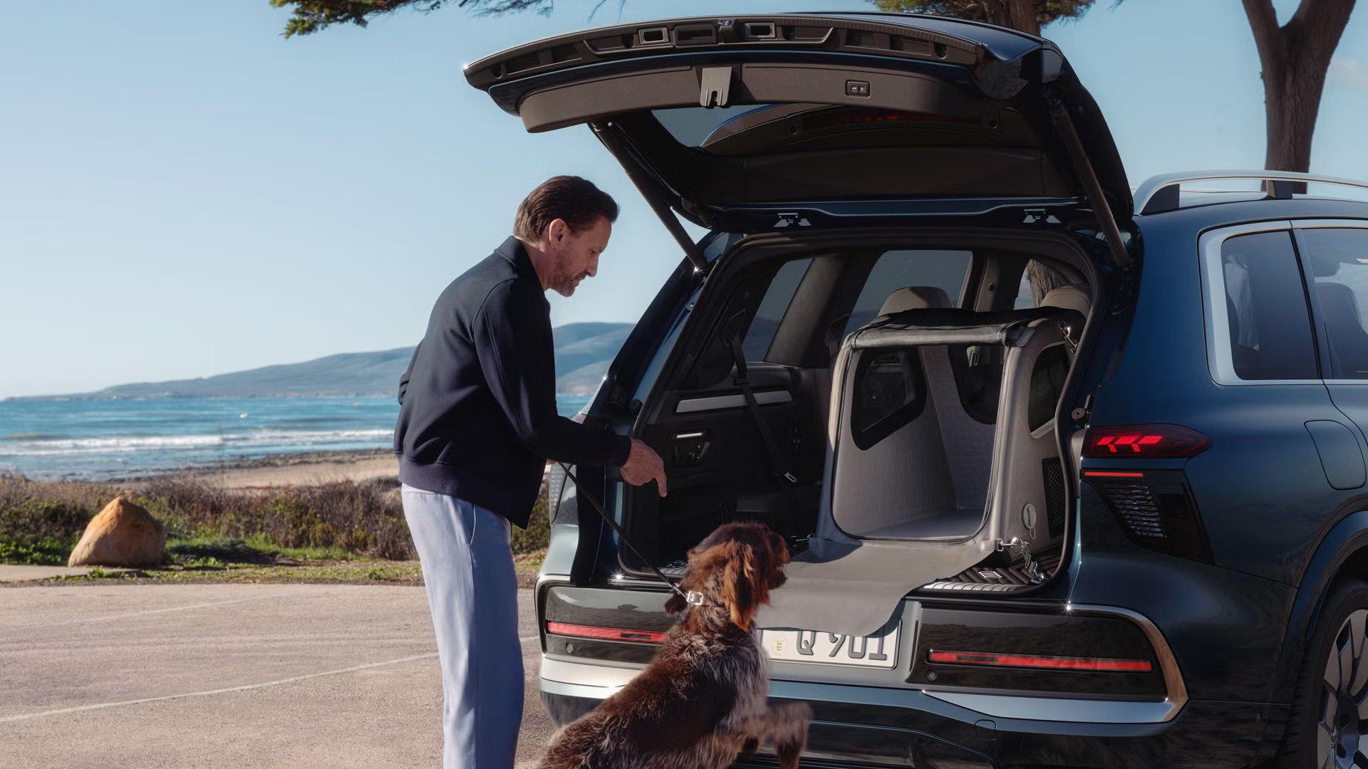 Audi Q9 estacionado junto à praia de bagageira aberta. Um homem encontra-se junto do carro a dar indicações ao cão para entrar dentro da bagageira.