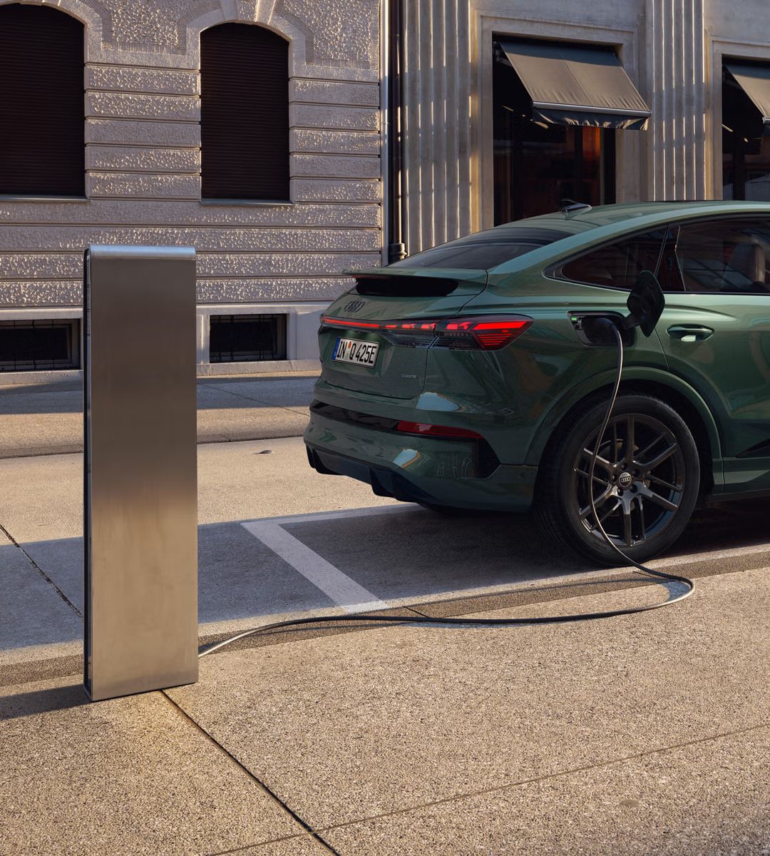 Blick auf das rechte Heck des Q4 Sportback e-tron, an einer Ladesäule parkend. Im Hintergrund ein modernes Gebäude. Das Kabel der Ladesäule ist mit dem Fahrzeug verbunden.