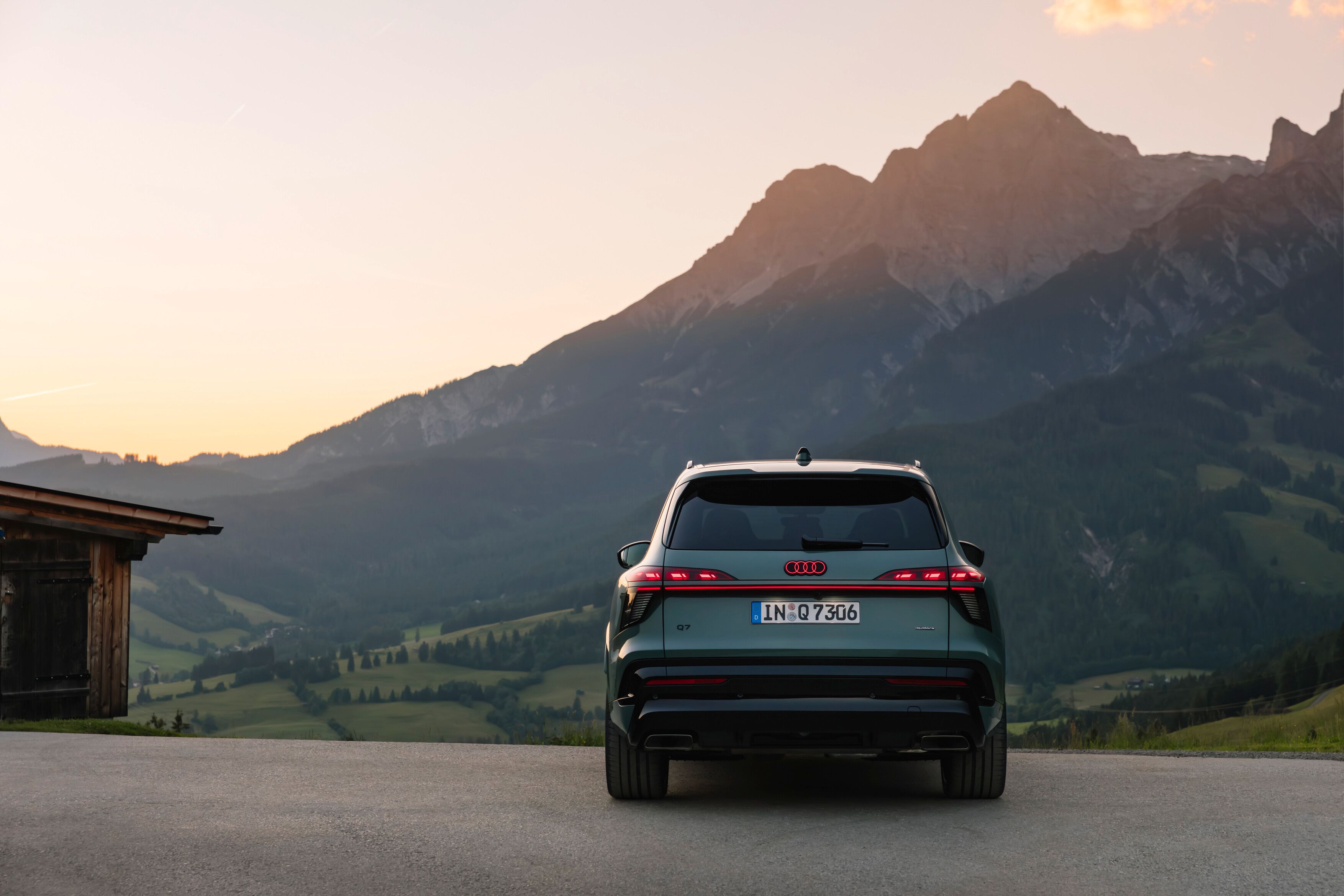 Vista traseira do novo audi q7 numa estrada com vista para um vale verde. Ao fundo, montanhas altas e rochosas destacam-se sob um céu de fim de tarde com tons suaves de laranja e azul.